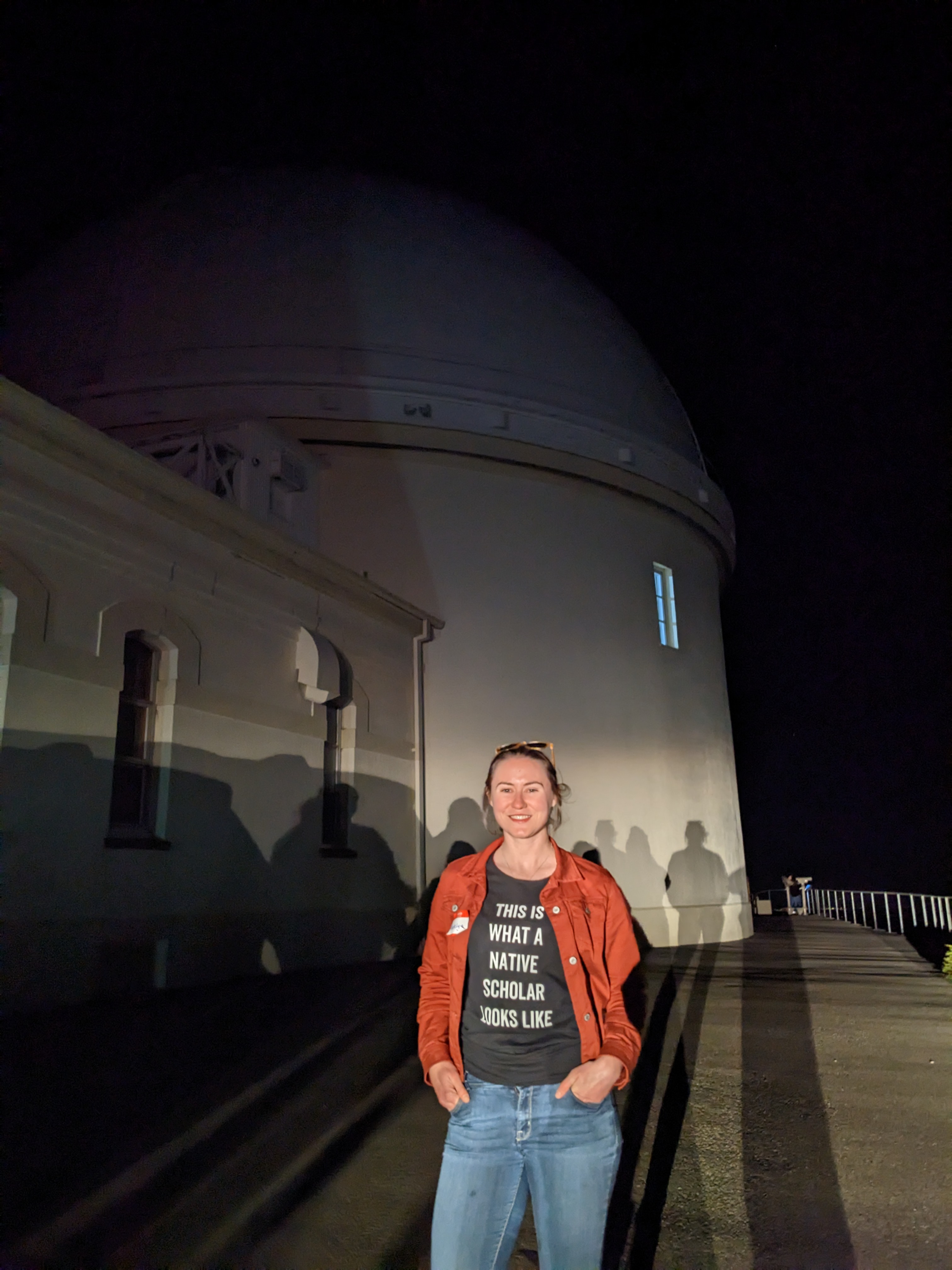 Madelyn stands in front of a Lick Observatory dome wearing a shirt that says, This is what a native scholar looks like.